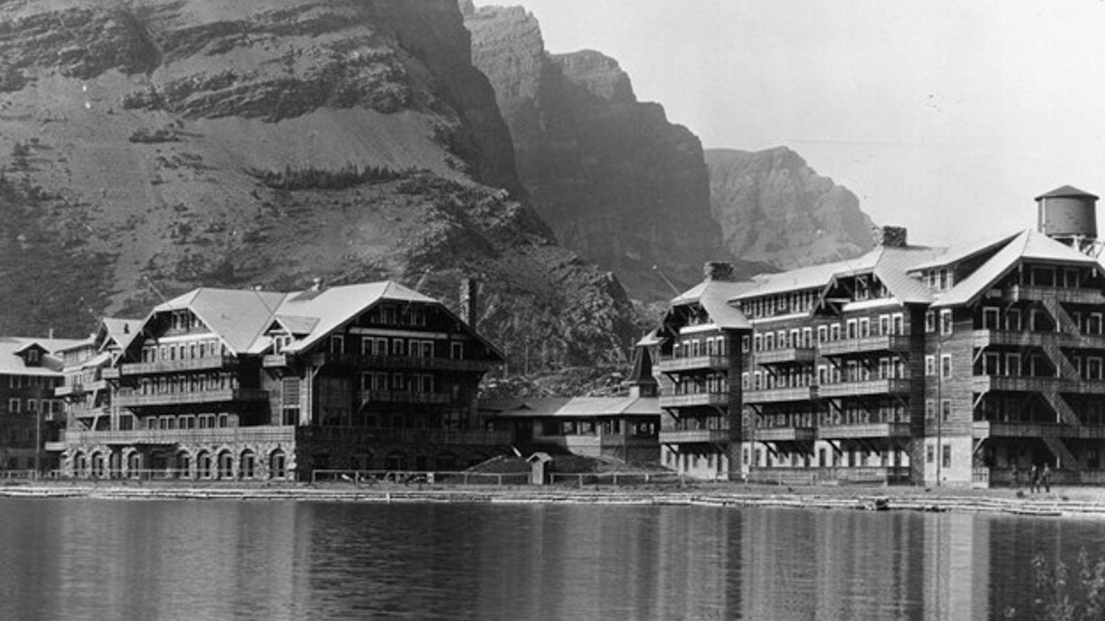 A glassy lake sits in front of a Swiss-style hotel and large mountains stand behind the hotel.