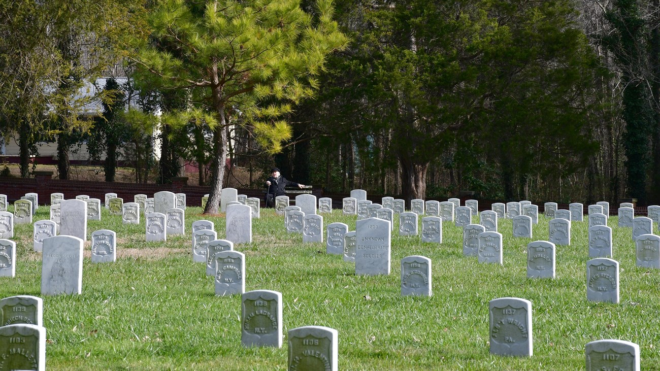 A man in a dark suit sits on a bench surrounded by white marble headstones.