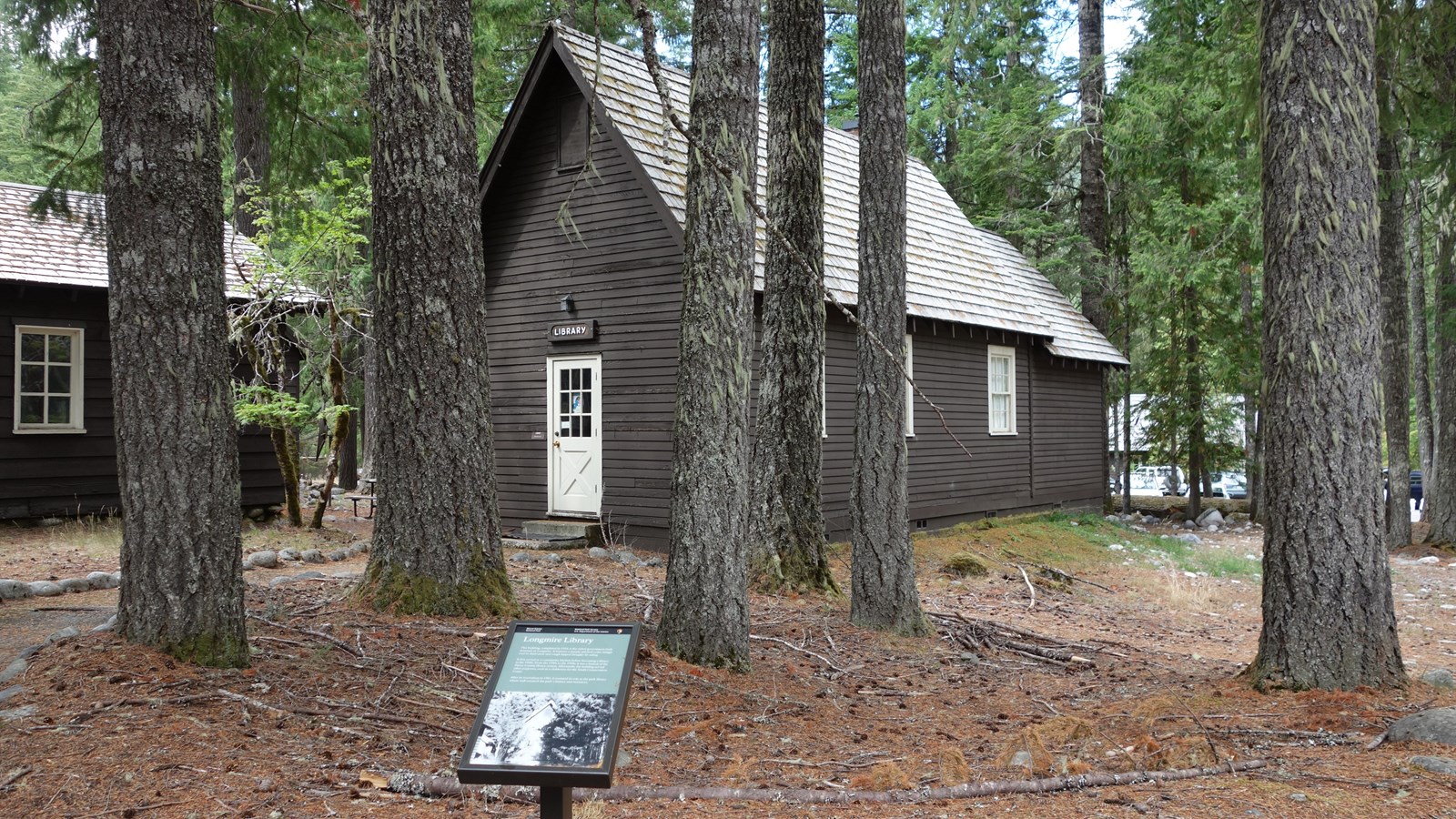 A small wayside panel in front of a narrow wood building with steeply angled roof.