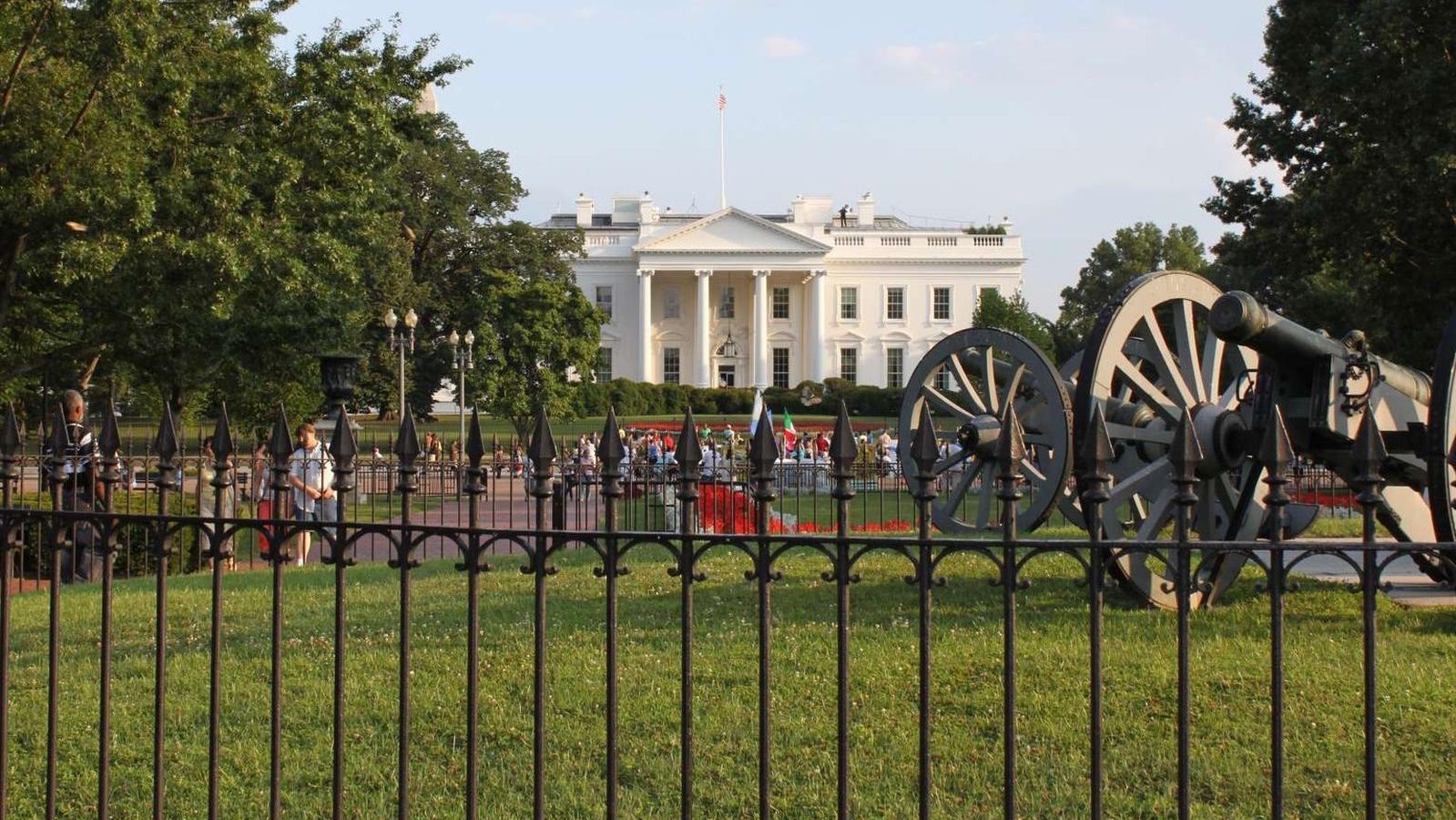 A cannon sits in a circular spot of grass surrounded by a fence. The White House is behind it.