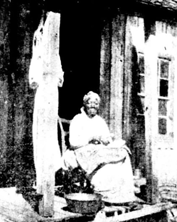 A black and white photo of a dark-skinned woman, in a long skirt sitting on the porch of a cabin. 