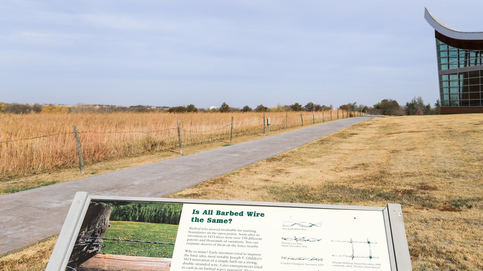 Informational sign stands in front of restored tallgrass prairie and a building shaped like a plow.