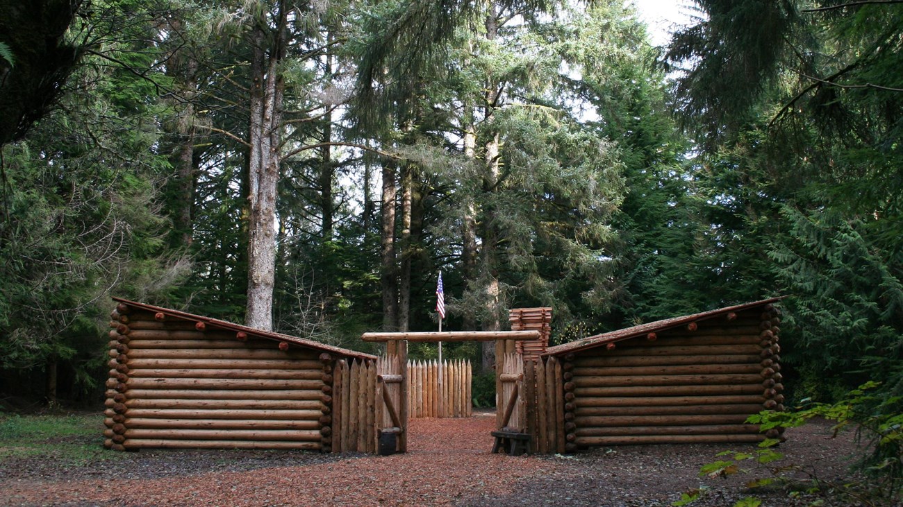 A log fort in a dense evergreen forest. Two parallel log structures with a flag in between.