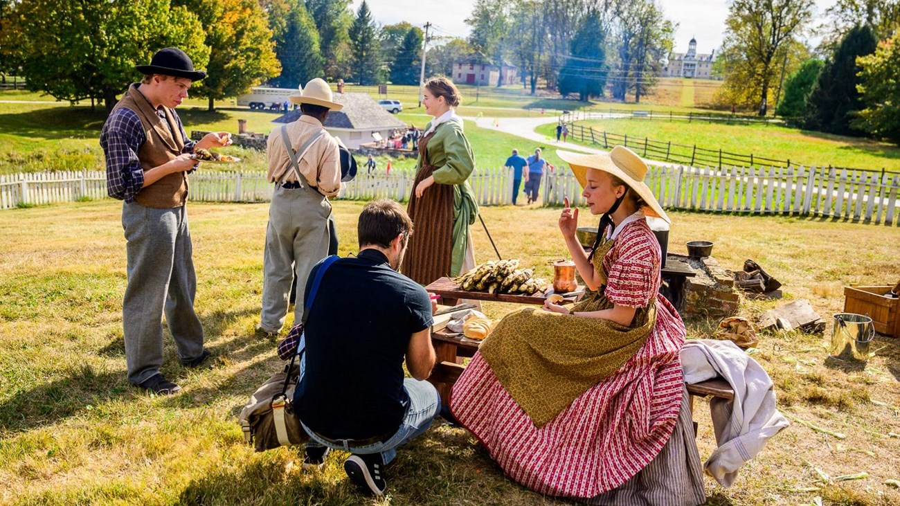 Living historians eating food at a wooden table on a sunny day.