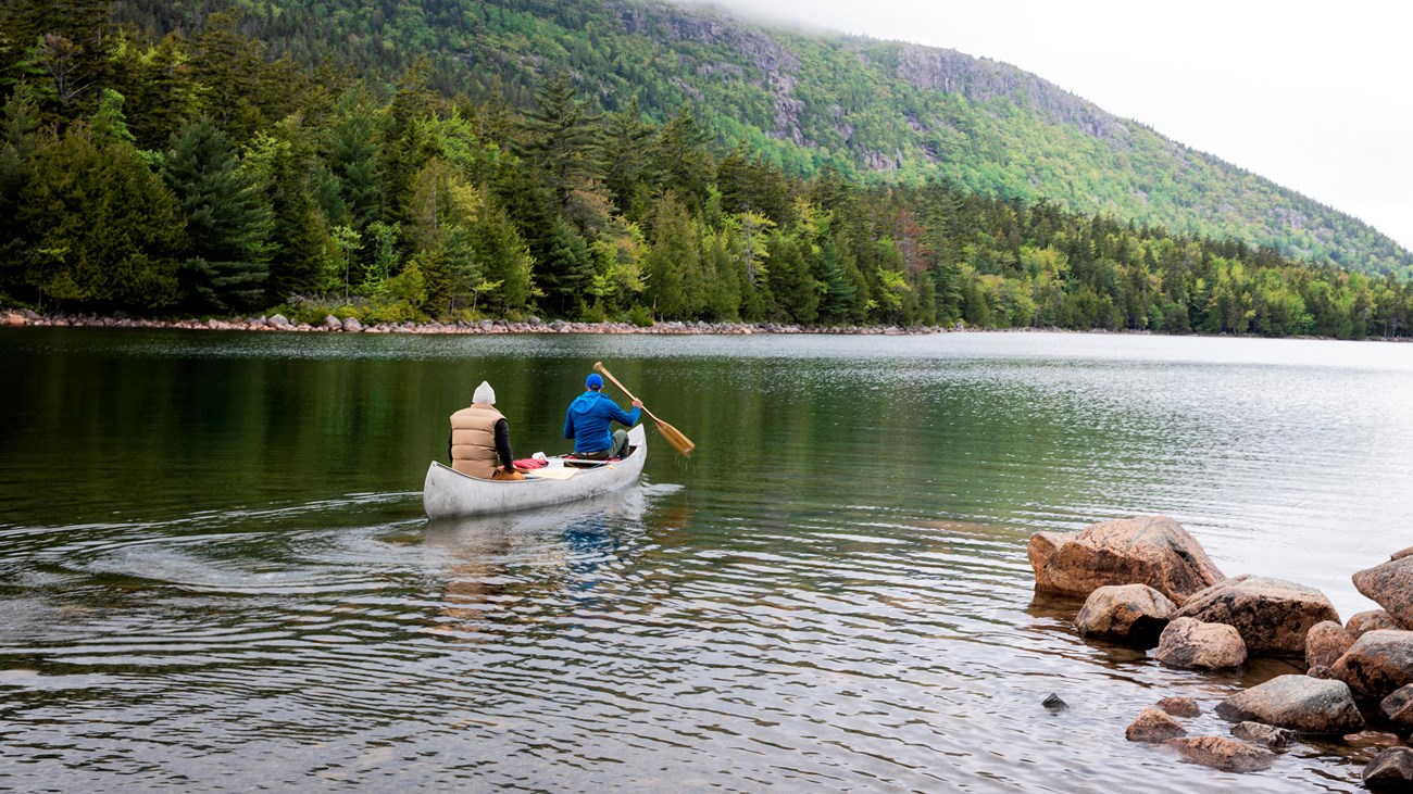 Two people, wearing jackets and hats, paddle a silver canoe in water surrounded by trees.