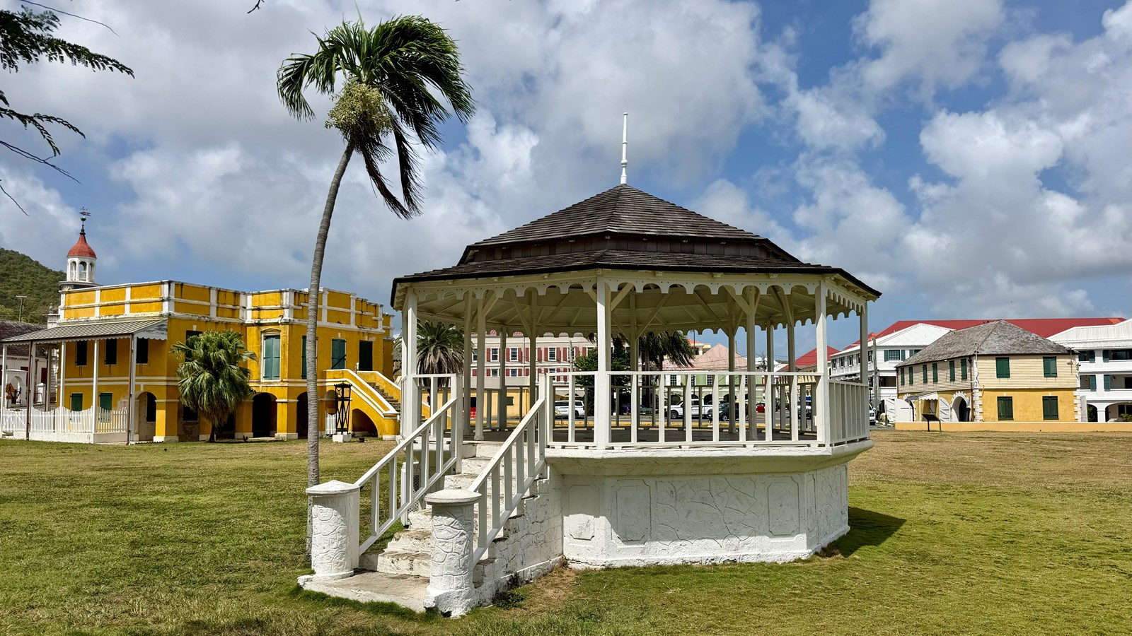 A white bandstand with colonial buildings in the distance