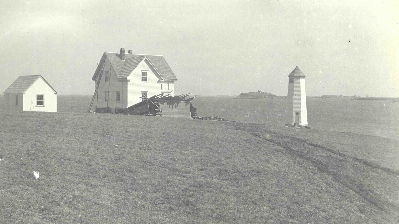 A white octagonal lighthouse next to two small white houses on an open grass area. 