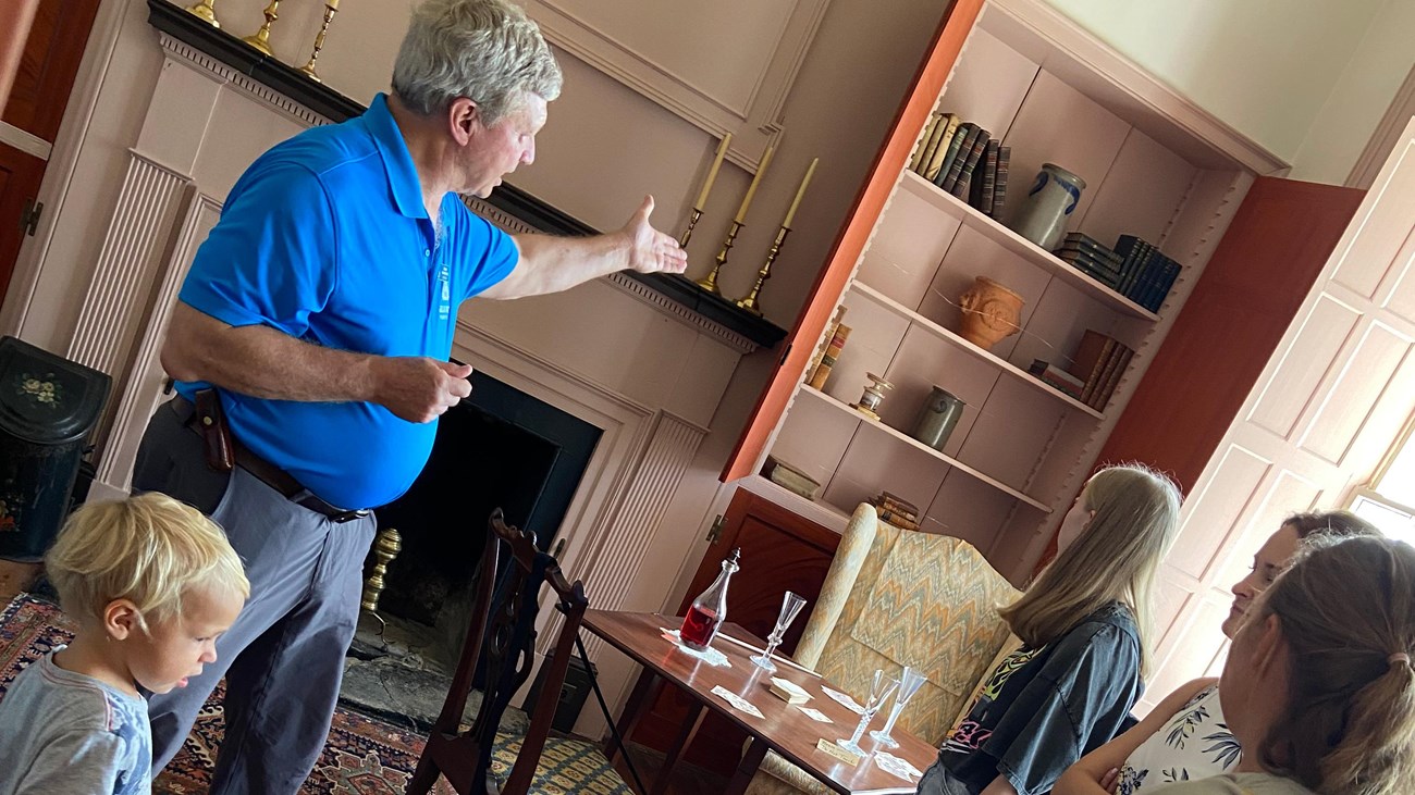 A man in a blue shirt leading a tour group gestures to items in a 1700s mansion.