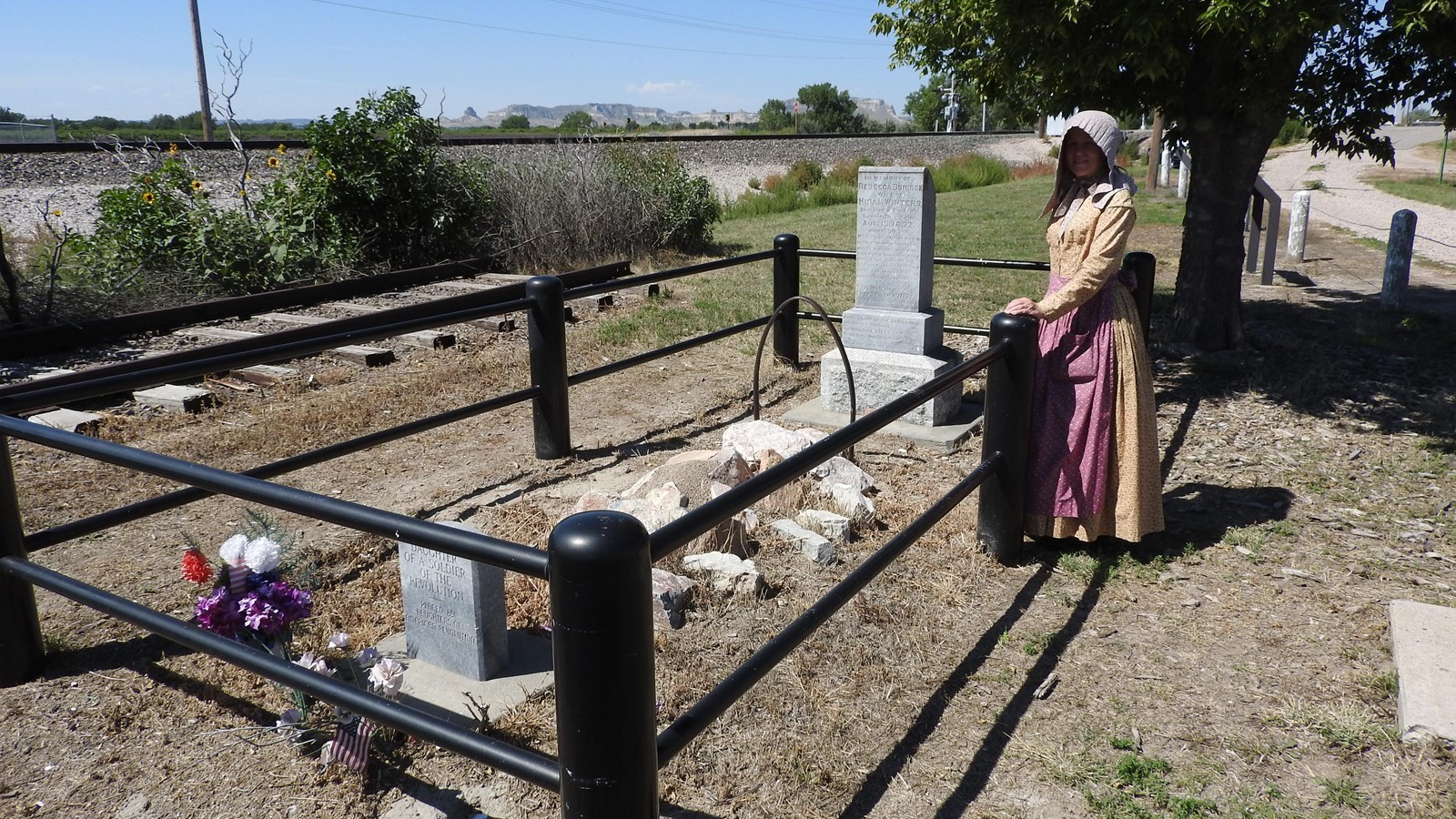 A woman dressed in emigrant costume is seen at the side of a grave.