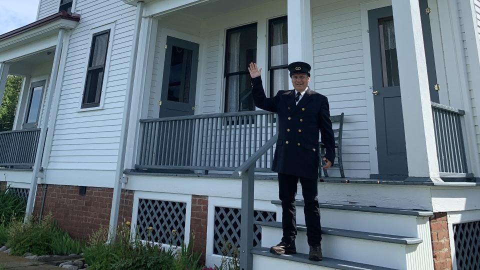A man dressed in black suit waives from porch steps of a two story white lighthouse.