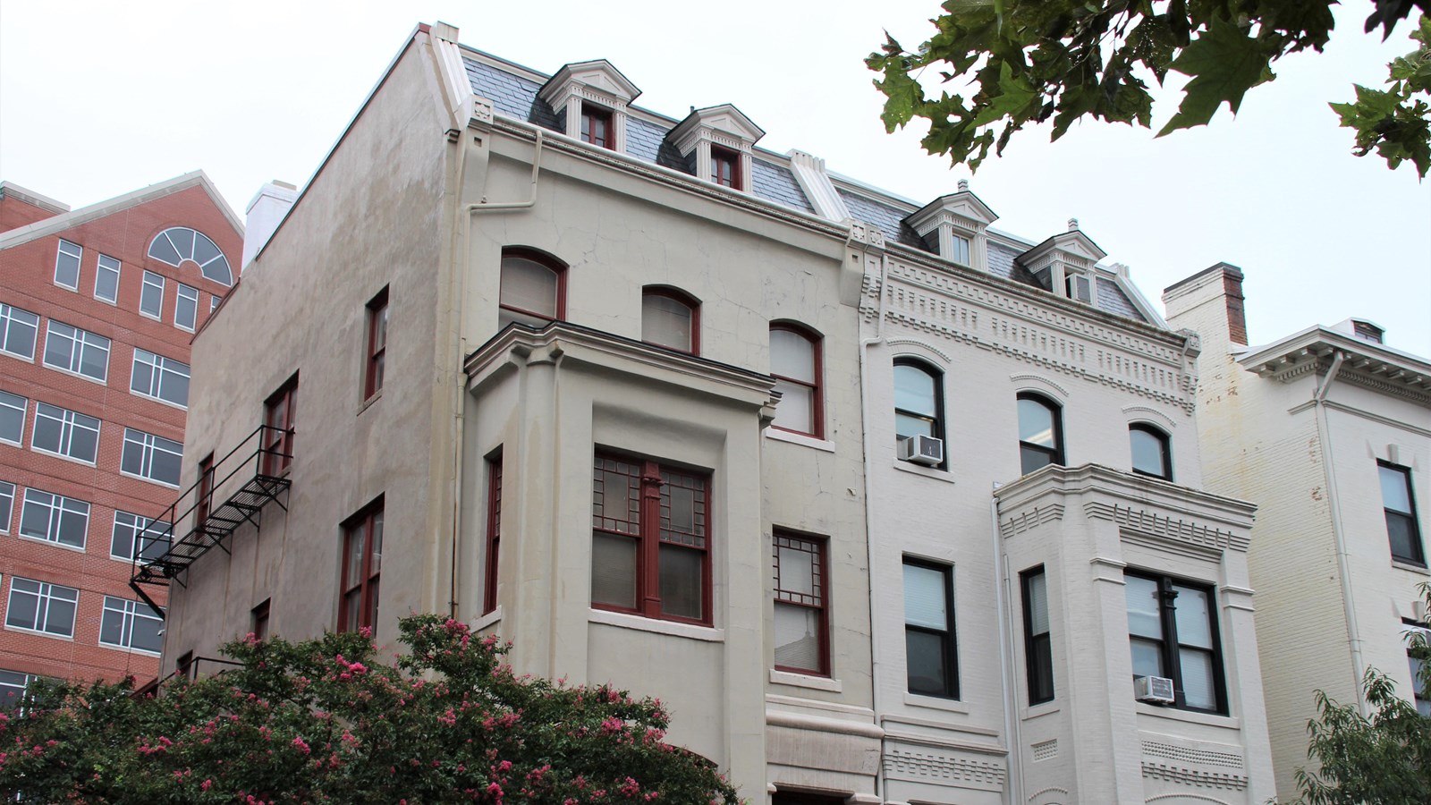 Three-story white plaster townhouse with red window frames and a bay window. 