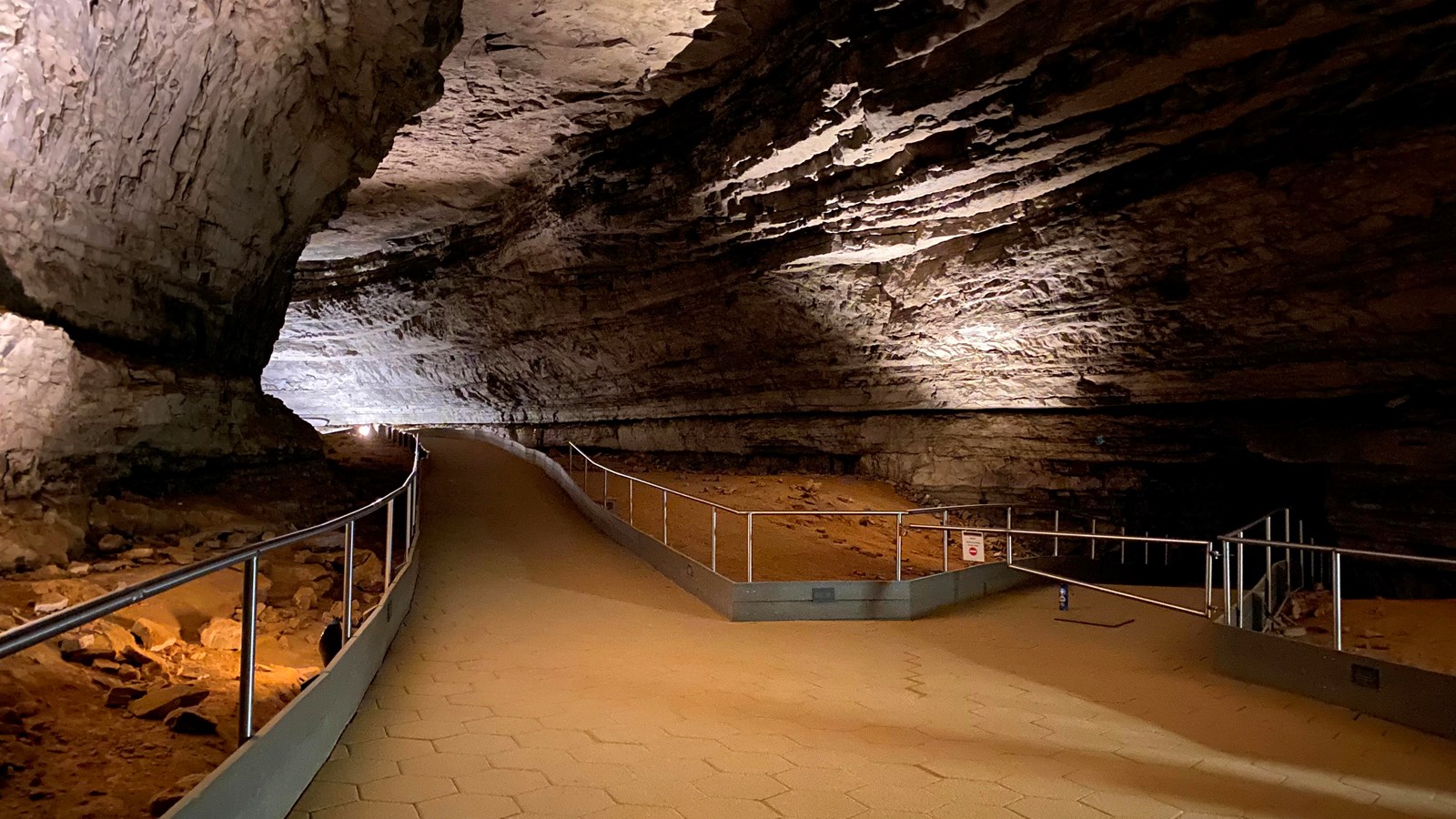 A large open cave passage with paved trails and metal handrails. A small dark passage leads away.