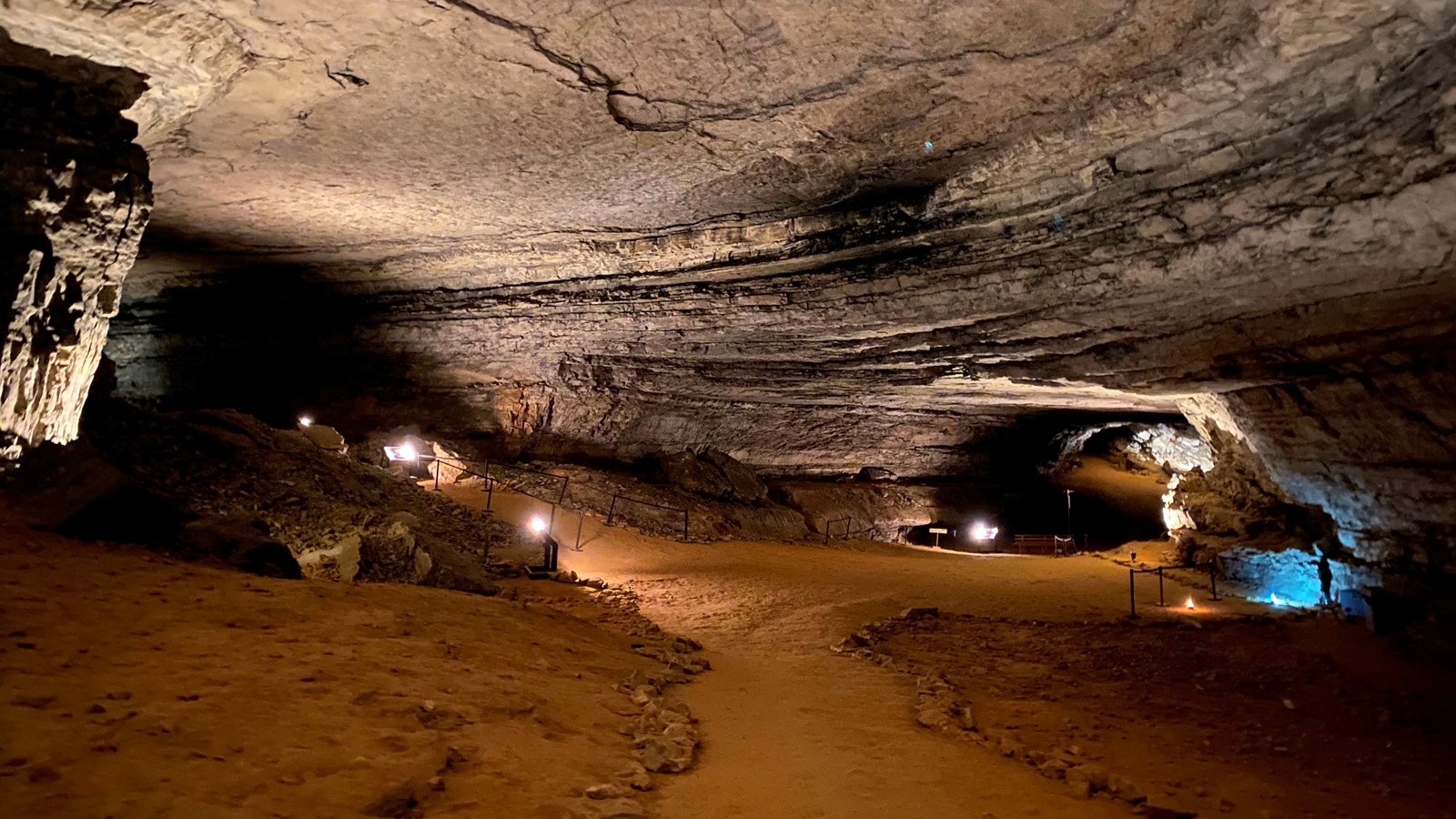 A large open cave room with dirt trails lined with rocks. 