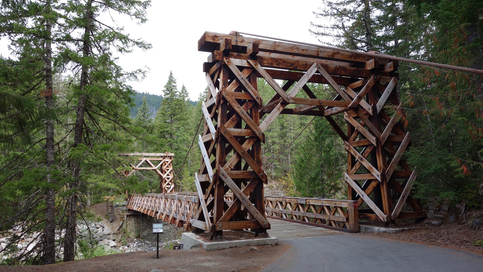 A small wayside next to a wood suspension bridge supported by stout wooden towers.