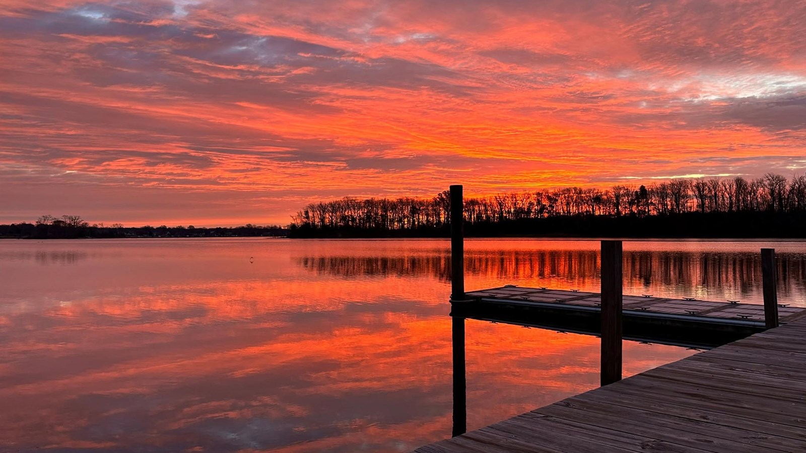 A dock extends into a river under a vivid orange and pink sunset, with reflections on calm water.