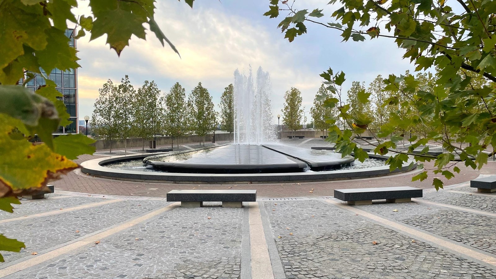 A fountain in the center of stone-clad plaza, with benches in front of the fountain.