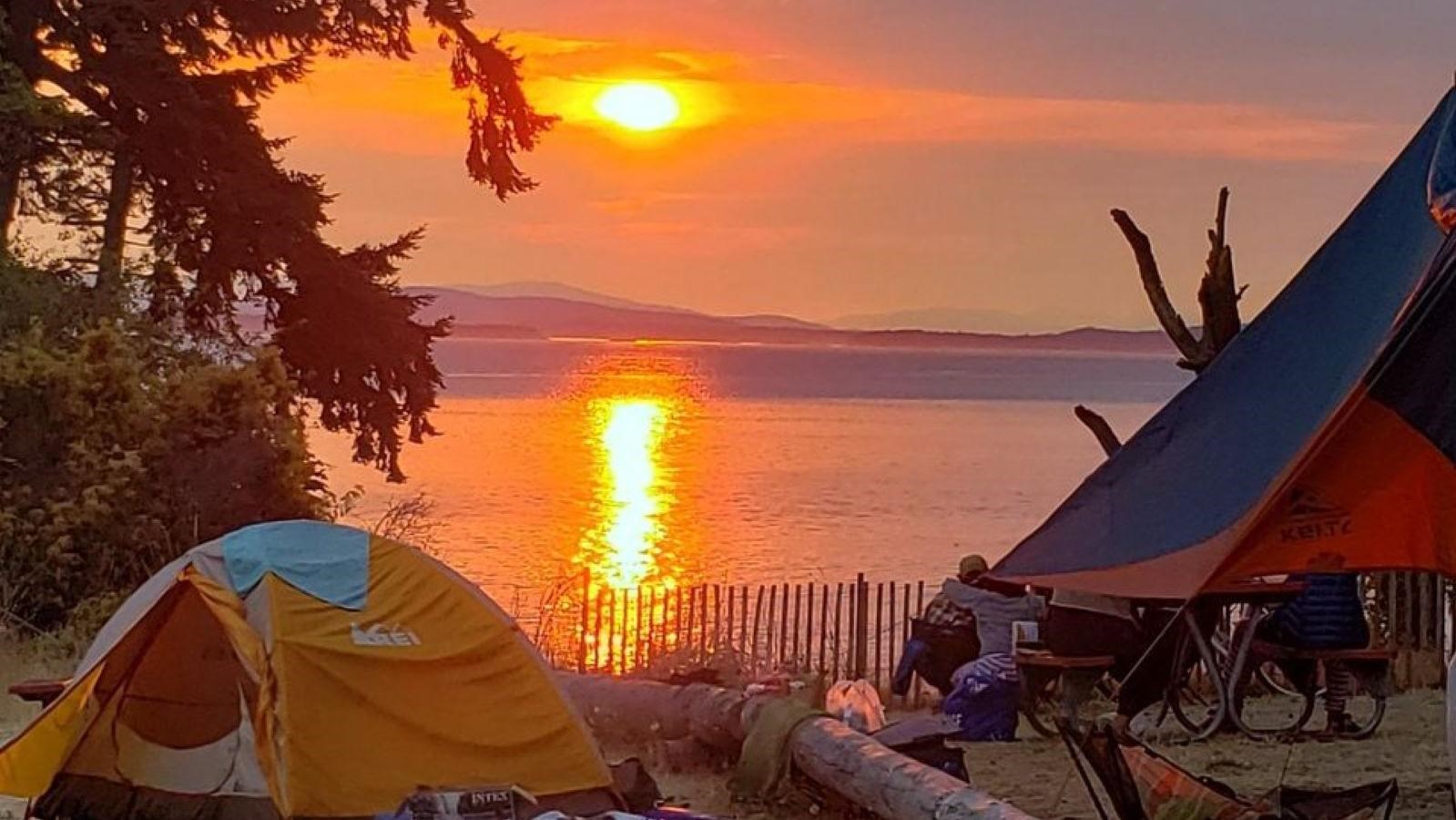 Sun setting over a waterfront campsite with a pitched tent and campers sitting at a picnic table