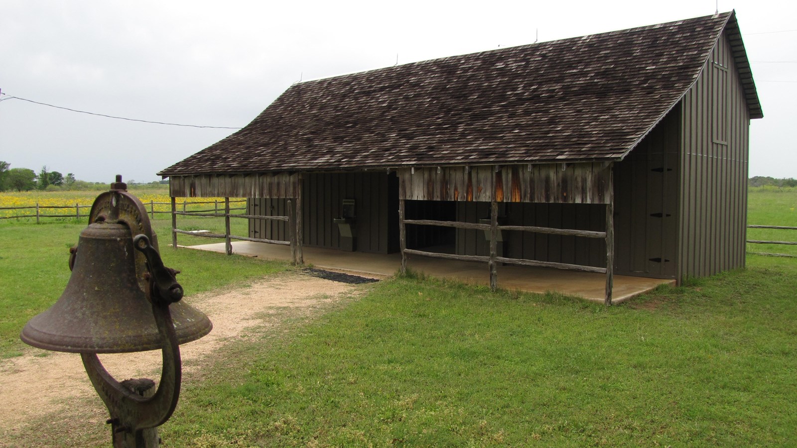Wooden barn-looking building surrounded by green grass stands behind a rusted bell on a post.