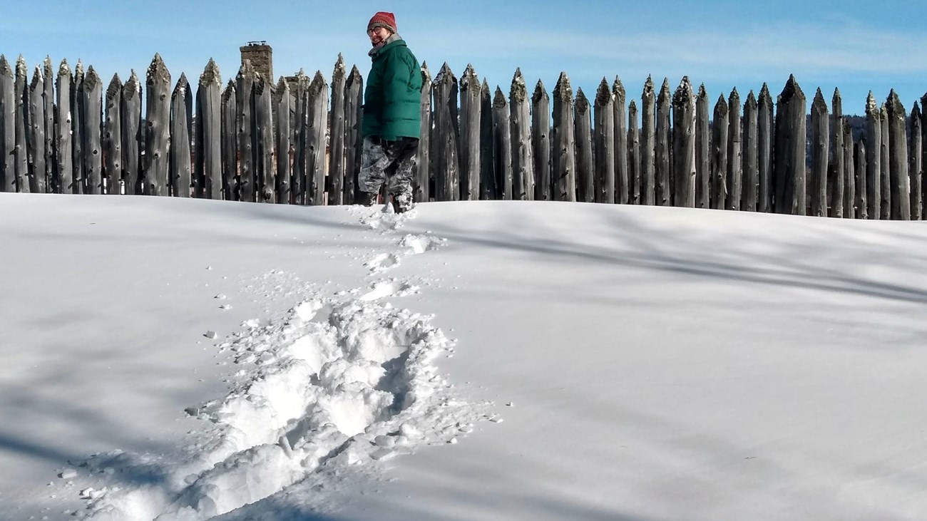 A person makes snowshoe tracks in front of a stockade.