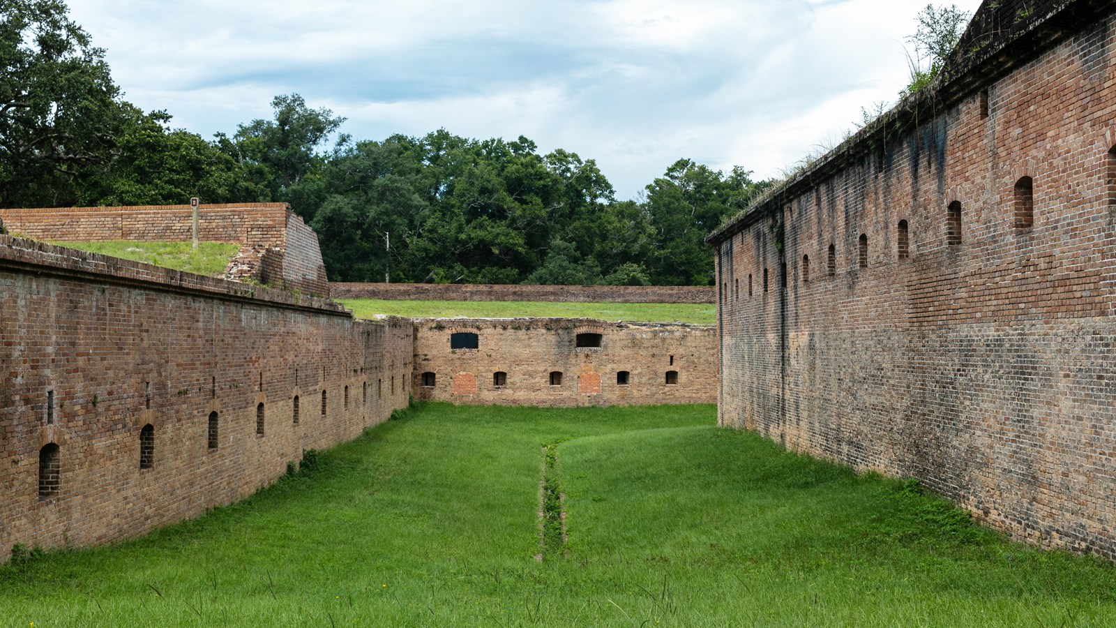 A grassy plain in between two brick walls of a fort leads to two closed off cannon embrasures.