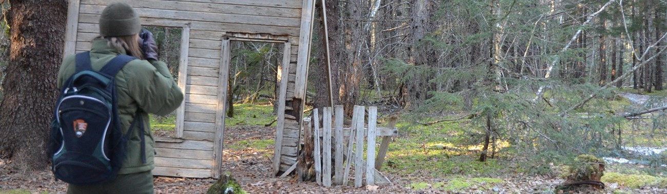 Woman in National Park Service uniform looking at remains of historic wooden building.
