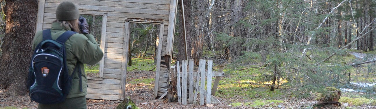 Woman in National Park Service uniform looking at remains of historic wooden building. 