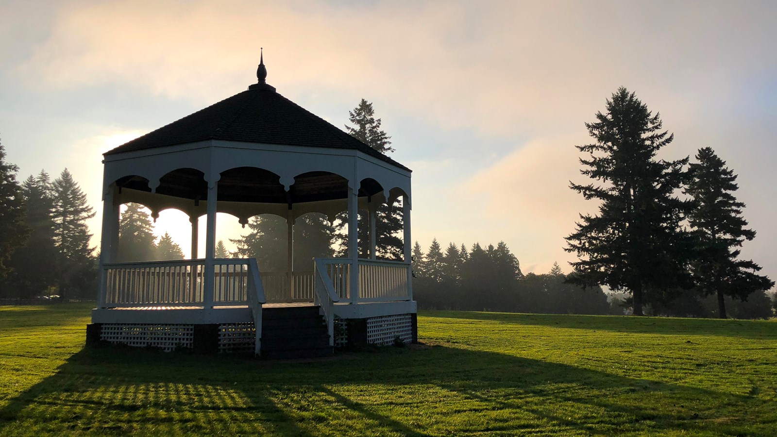 A white bandstand in a grassy field at sunrise.