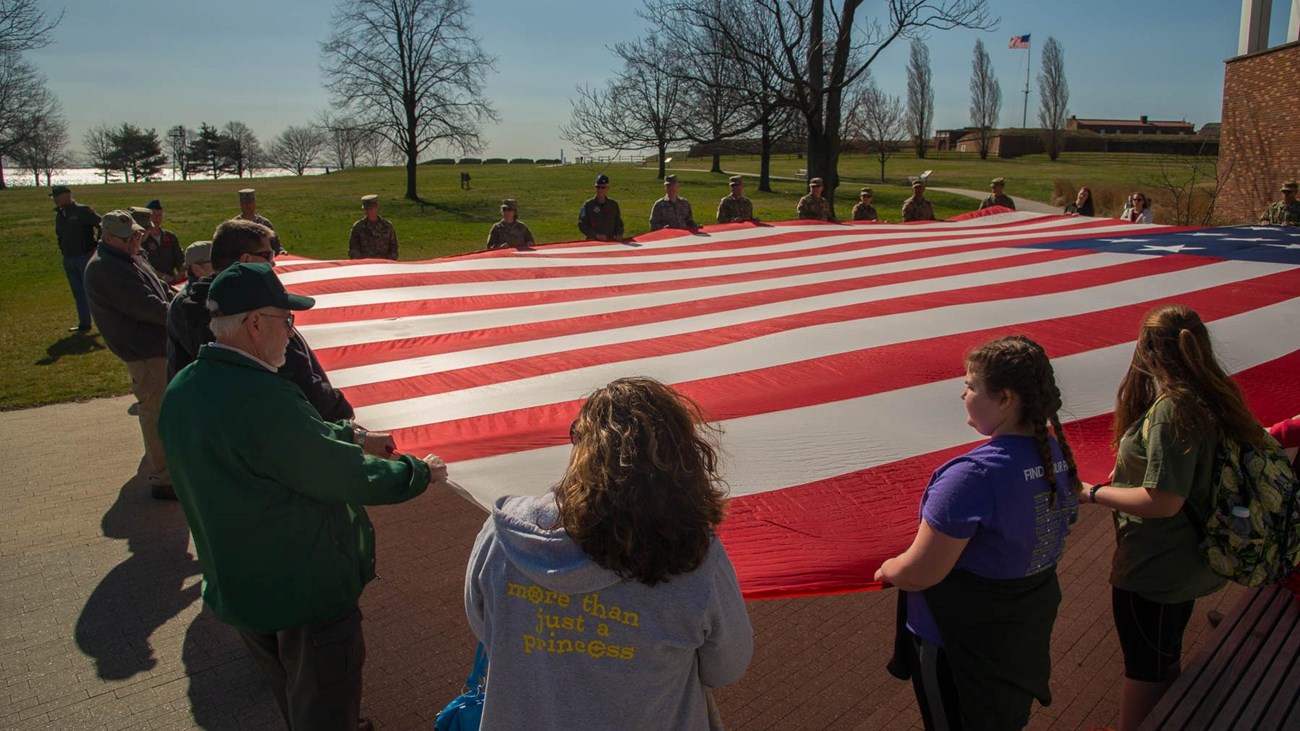 A group of visitors holding on to a large Star-Spangled Banner Flag.