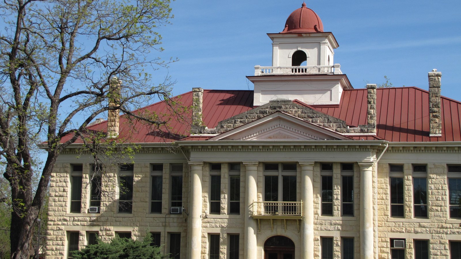 Under a blue sky, a large, limestone building has four center columns, a red roof and a domed cupola