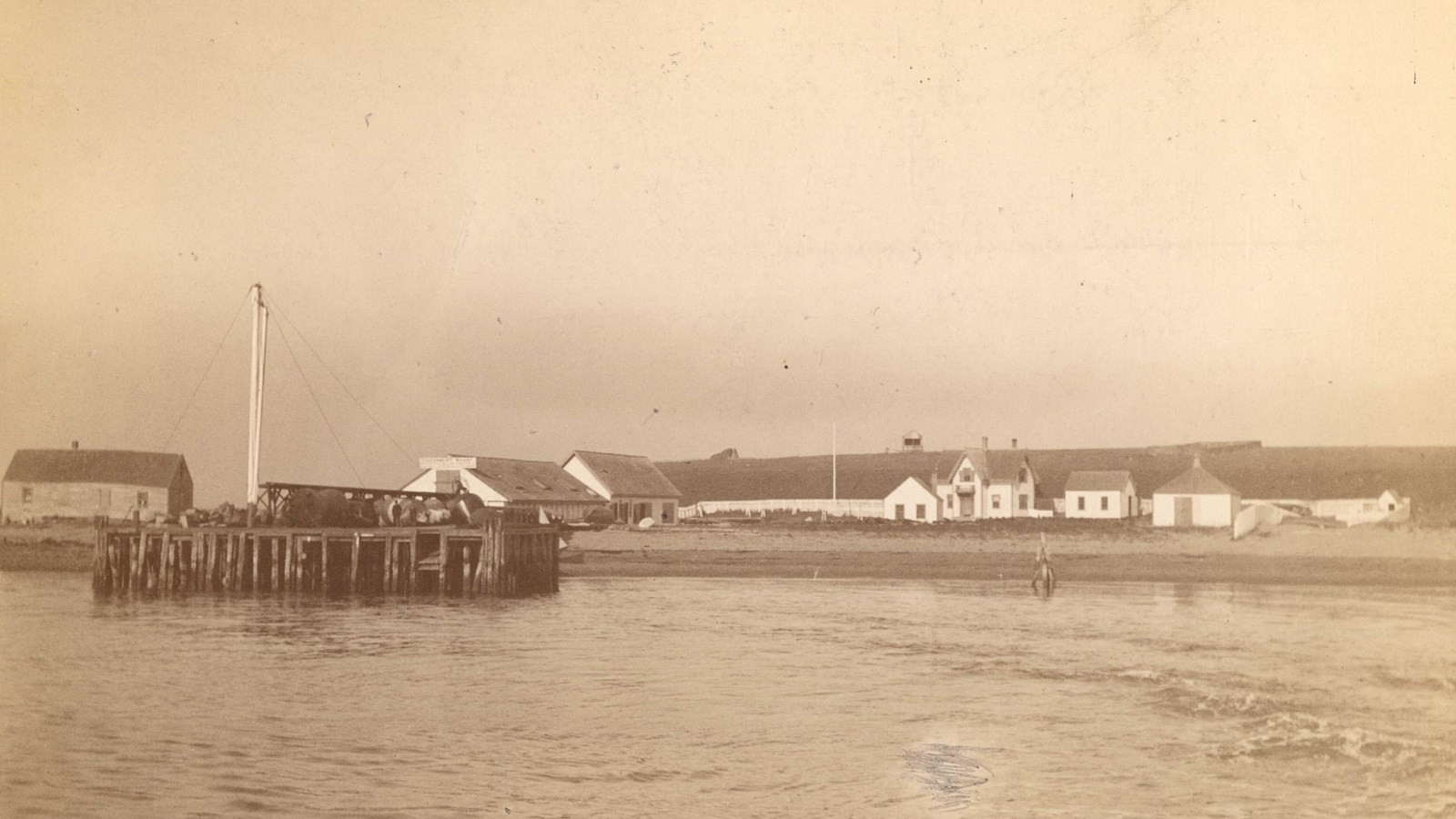 Sepia image of a shore with a cropping of white houses, with a prominent wharf in front. 
