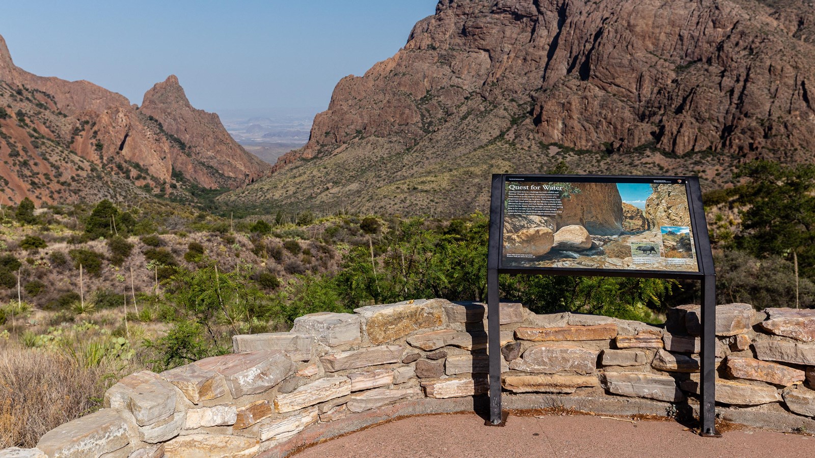 A paved trail and metal exhibit sign overlook a low basin surrounded by mountains.