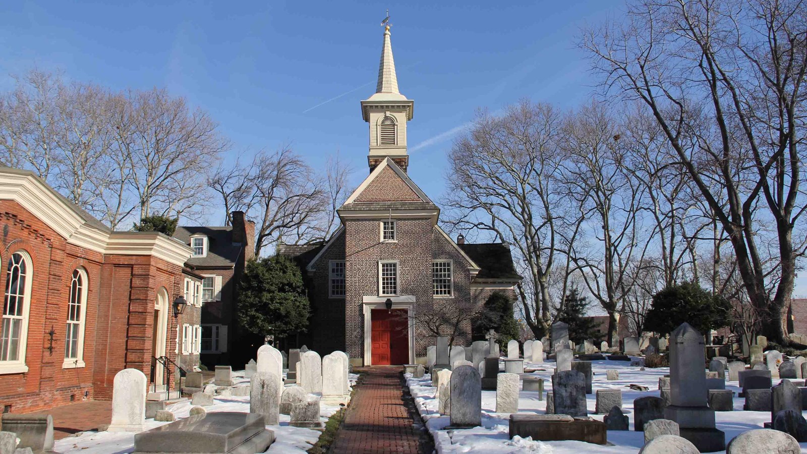 A brick church with a red door and wooden steeple behind a snow covered burial ground.