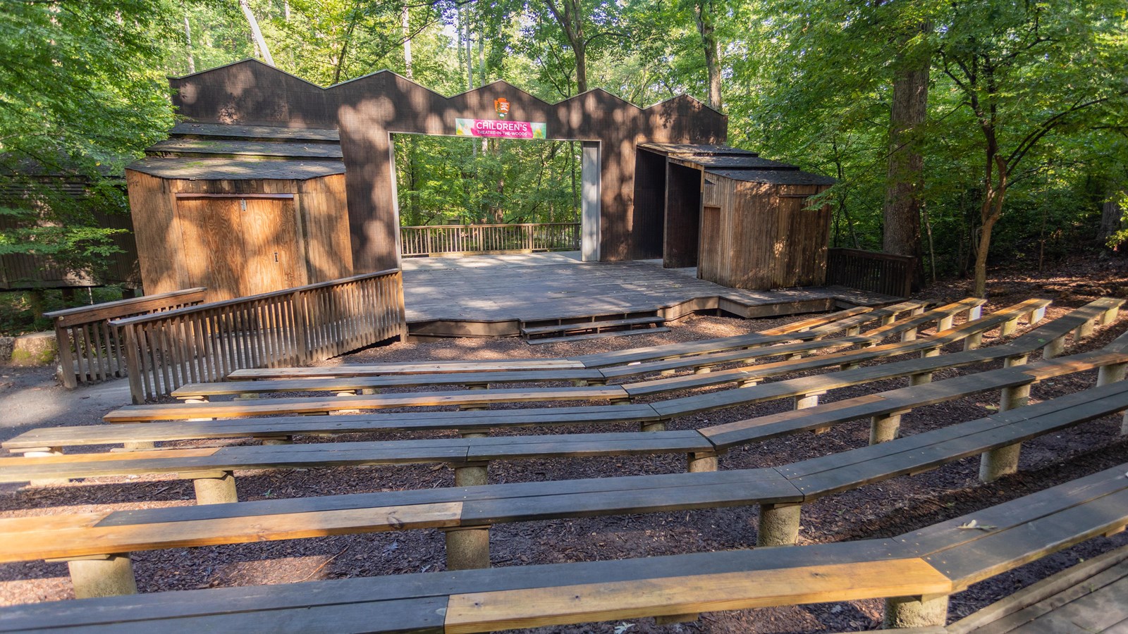 Wooden deck stage with bench seating in the shade of surrounding trees.