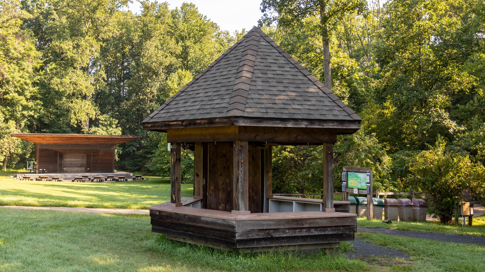 Wooden kiosk surrounded by grass with a stage area and trees visible in the background.