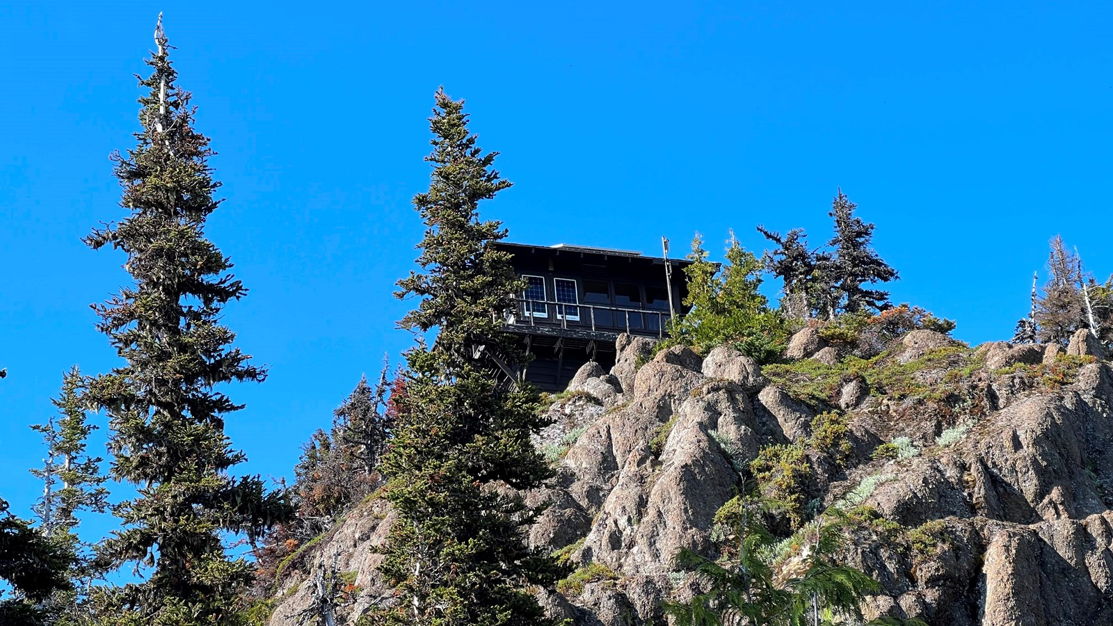A wooden fire lookout sits upon rocks with conifer trees surrounding it
