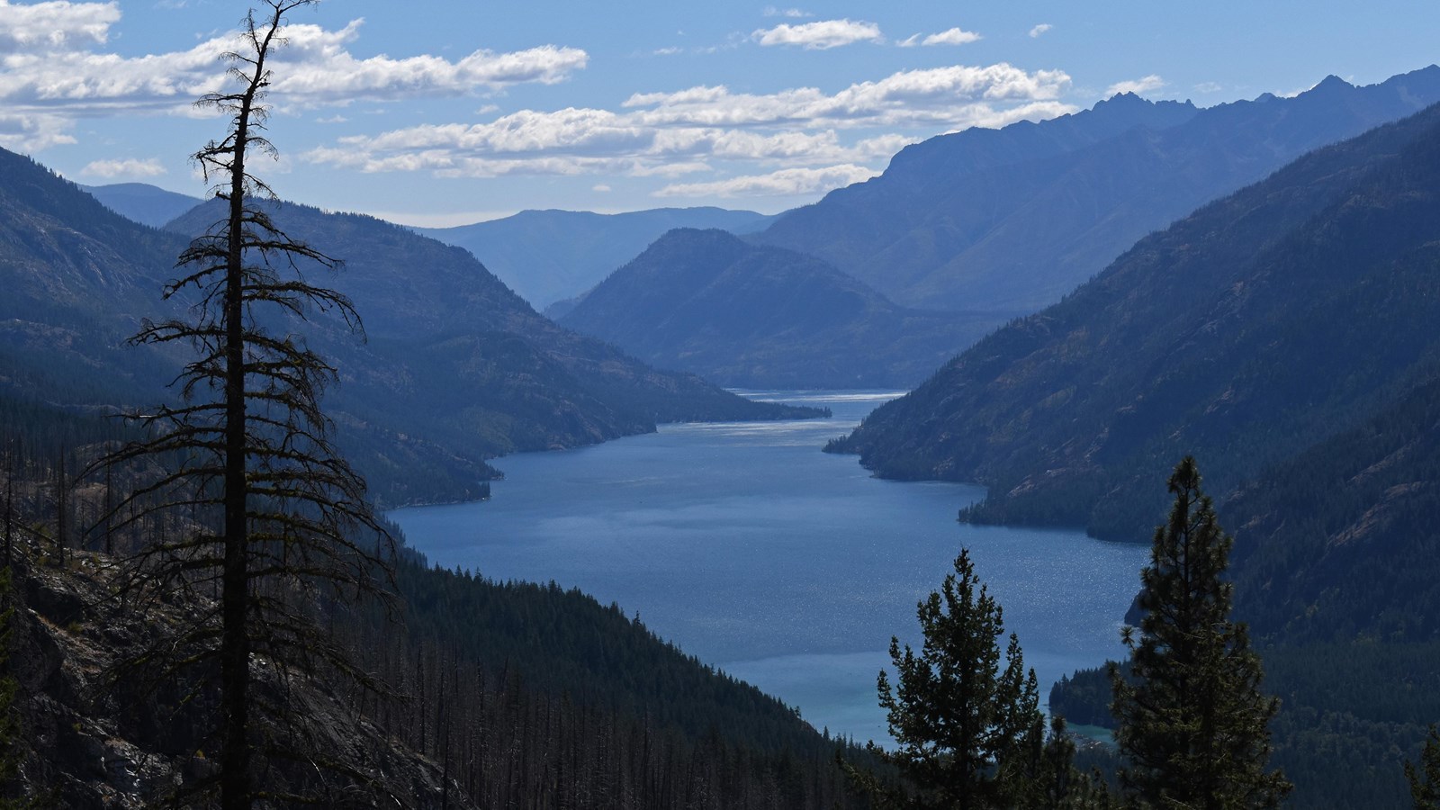 A view from elevation looking down on a long blue lake with steep forested hills on both sides.