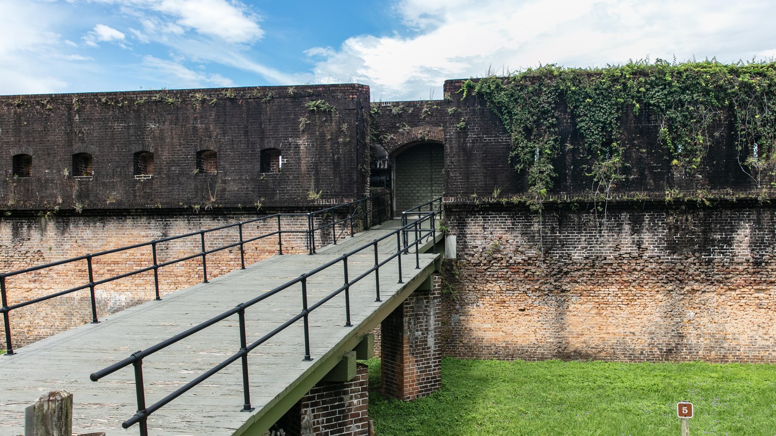 A drawbridge leads to the large doors of a fort with brick walls on either side.