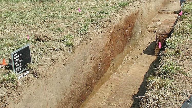 An archeological excavation trench shows red soil piled on yellow soil. Cars and trees in background