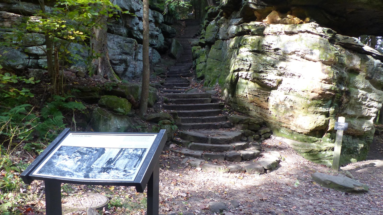 Gray stone staircase between two uneven rock walls; informational sign in brown metal frame at left.