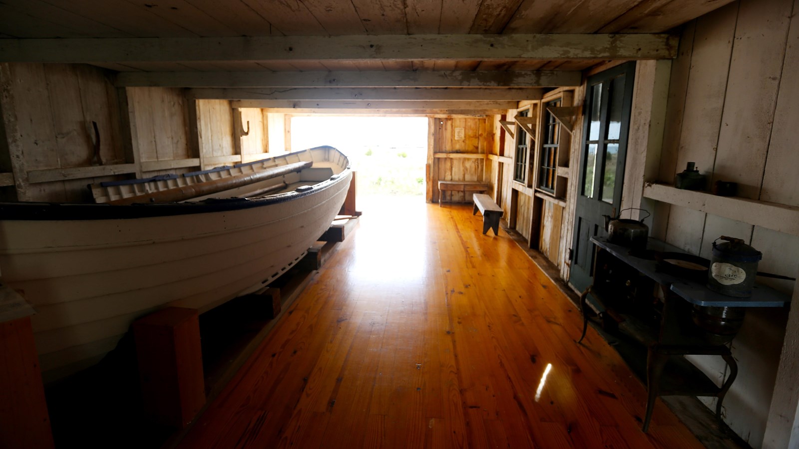 Interior view with wood floor, white rowboat, wooden benches, large open door, antique stove