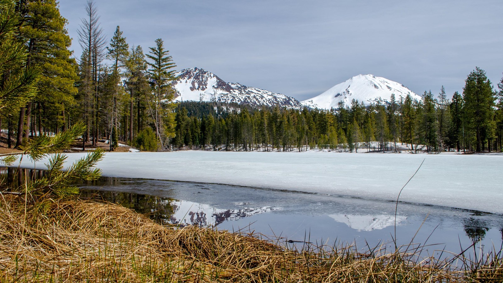 Two snow-covered volcanoes reflected in a small patch of unfrozen water backed by a frozen lake line