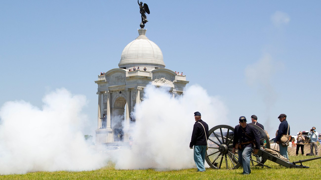 Living history volunteers fire a civil war cannon in front of a white, domed monument.