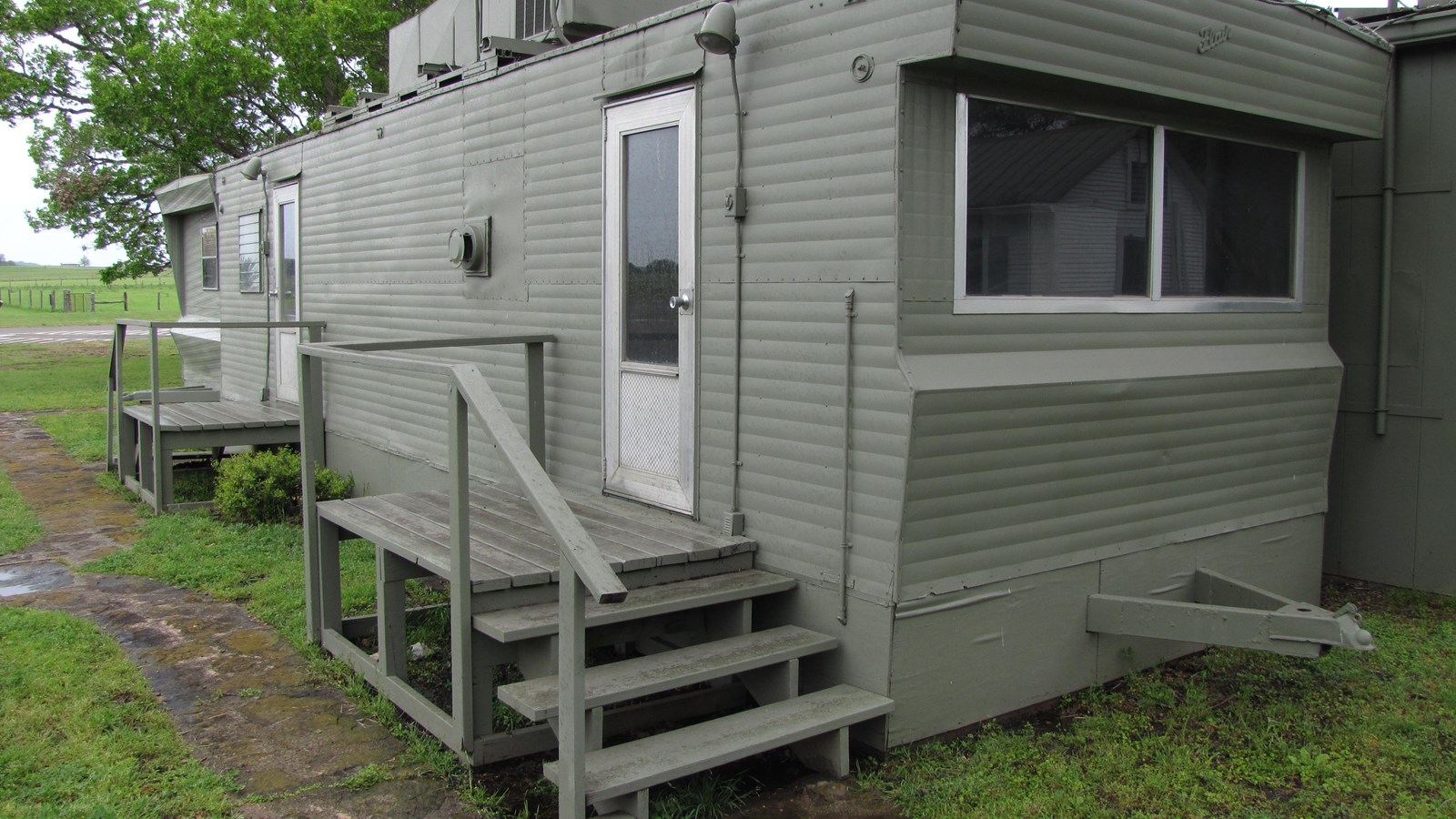 A long, jungle green trailer has a window at its end and two doors on its side with wooden staris.
