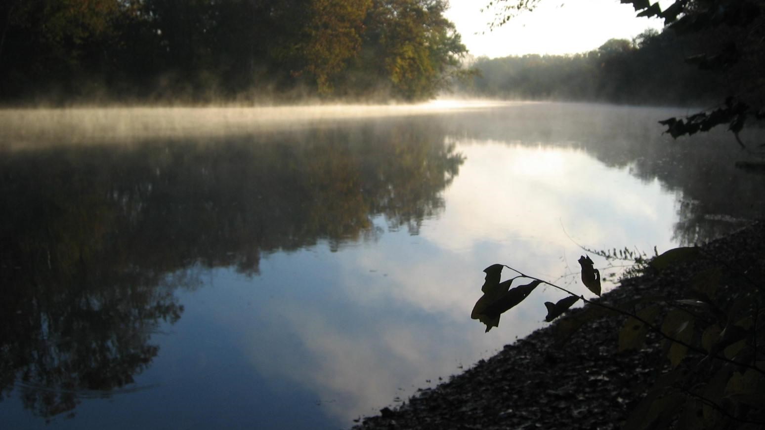 A color photo of a river at dusk, the banks can be seen on both sides with trees and gravel bar.