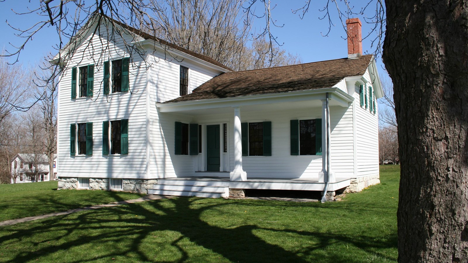 A two-story white house with green shutters, a low porch, and a large yard. behind.