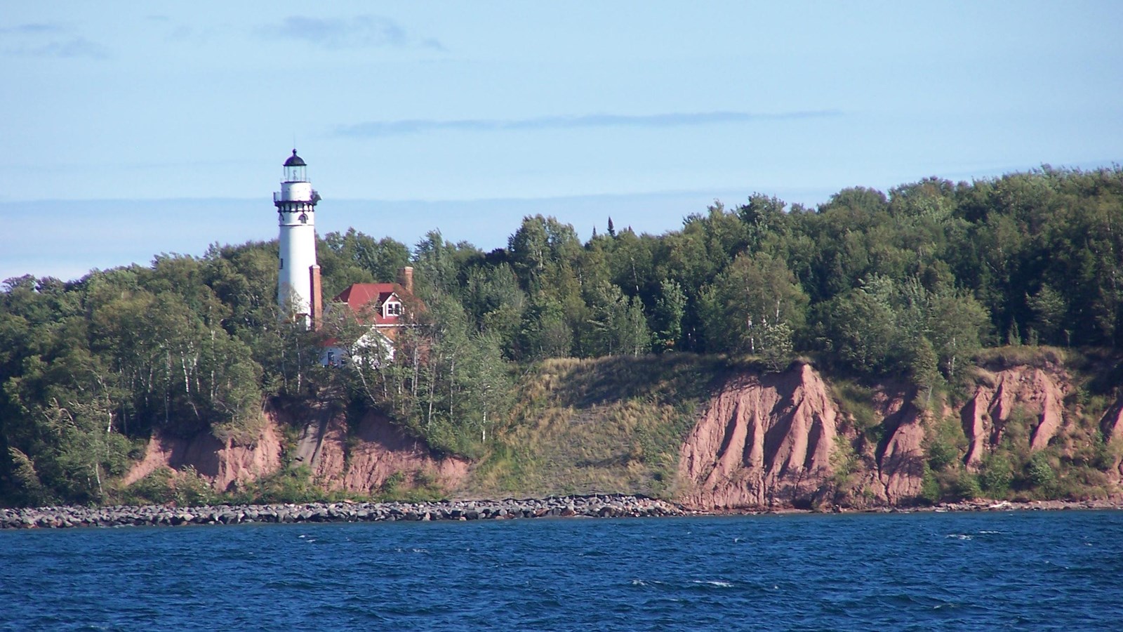 A white lighthouse tower stands tall on a steep clay bank with trees on the edge of a lake. 