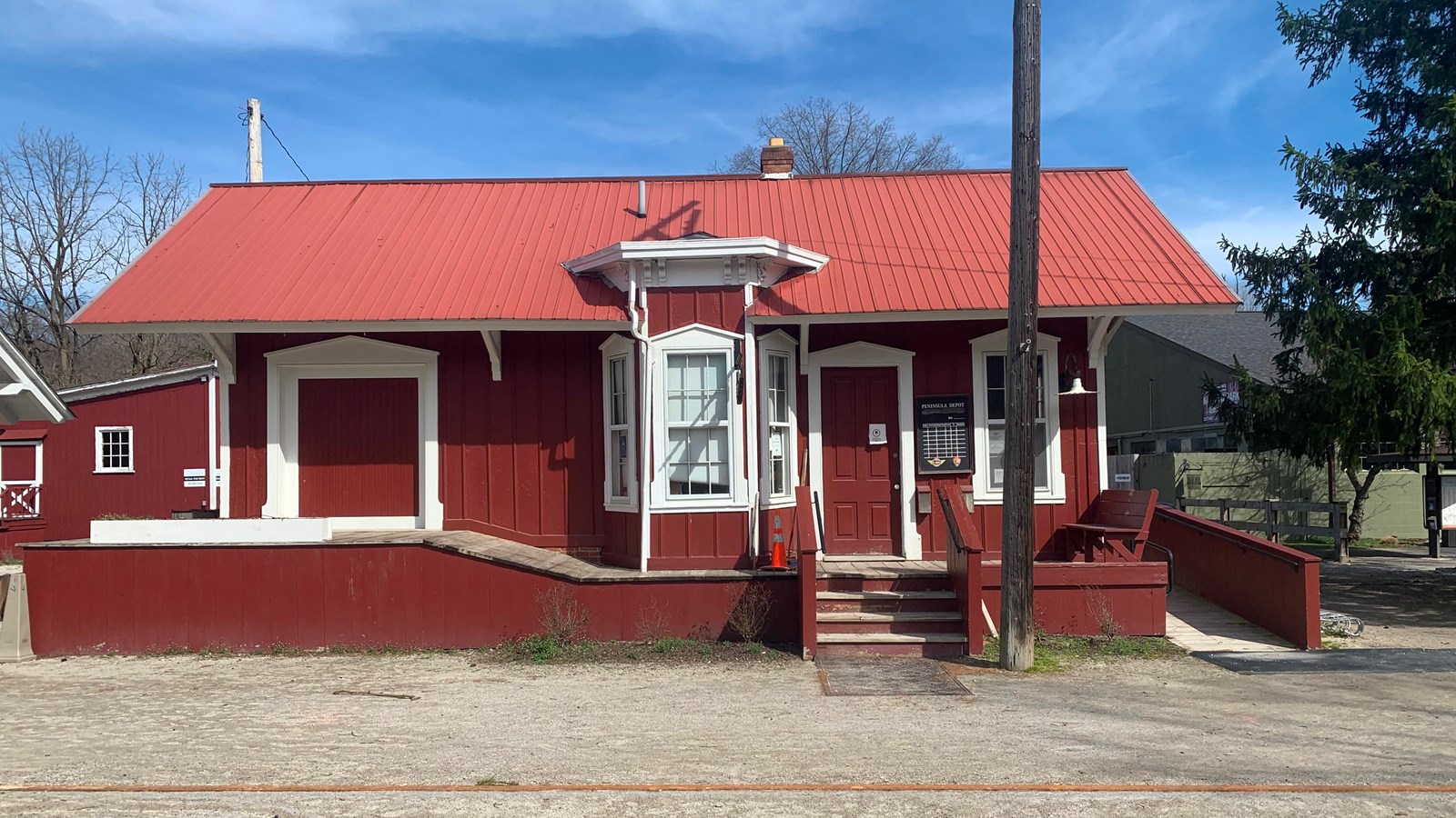 A red building with white trim. Depot has a platform, ramp, posted train schedule, and bay window.