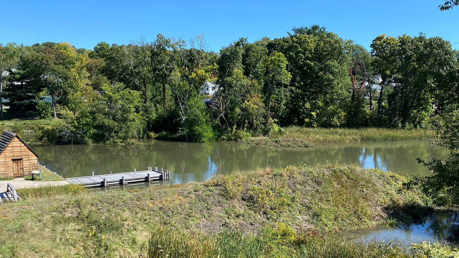 raised earthen mound covered in grass on riverbank across from small wooden warehouse and dock