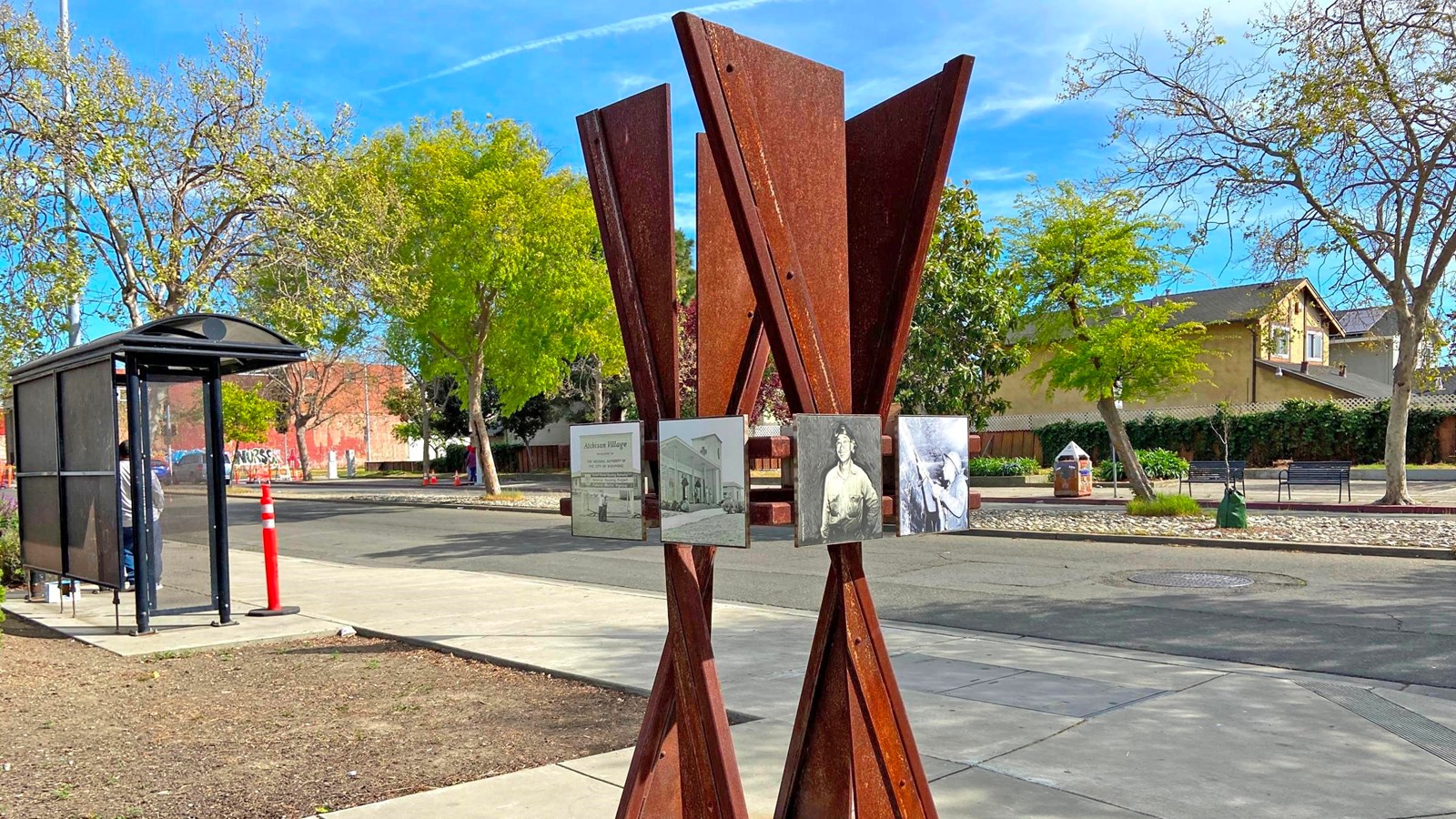 A metal sculpture with embedded photos, sits on a sidewalk next to the street.