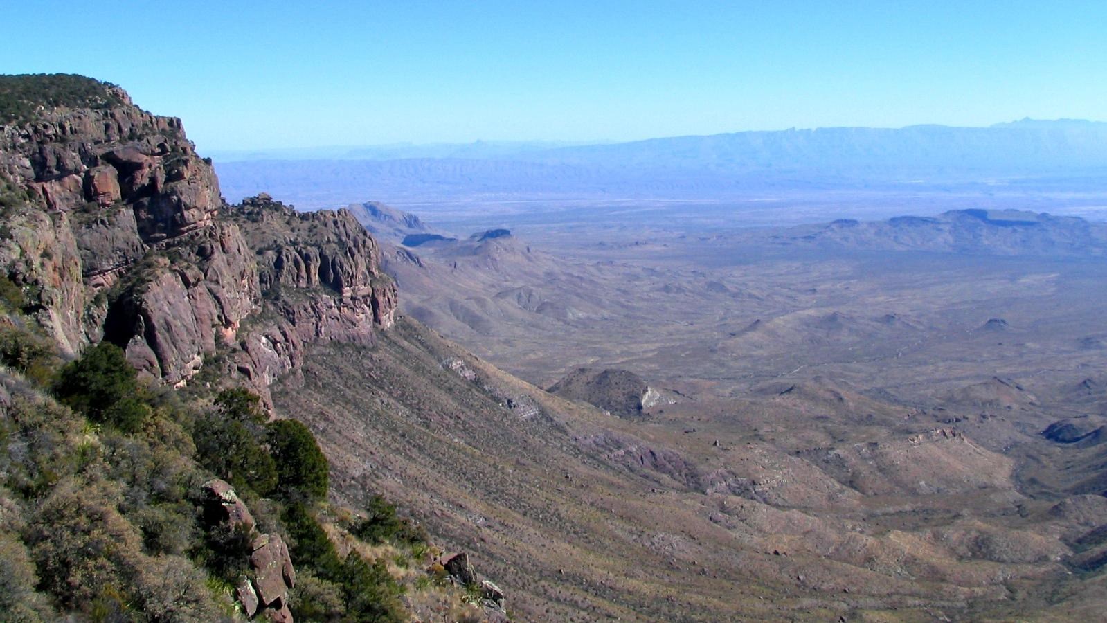 A view from the edge of a high cliff, looking down onto lower deserts and far away mountains.